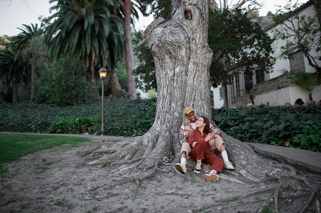 Pareja trans tomándose de la mano y mirándose a los ojos mientras están sentados junto a un gran árbol en el parque