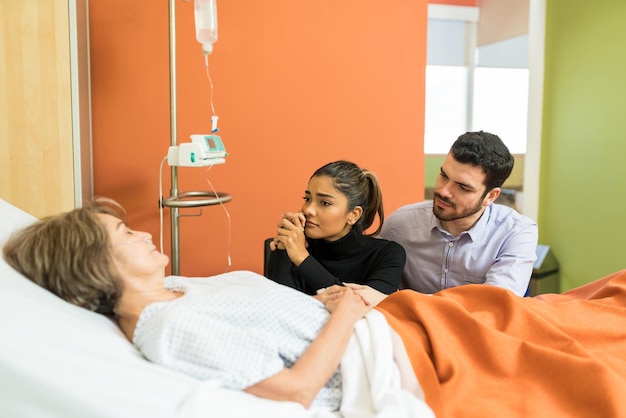 Una pareja triste mirando a una madre mayor sentada junto a la cama en el hospital