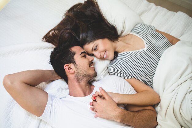 Pareja tumbada en la cama agarrada de la mano y mirándose a los ojos