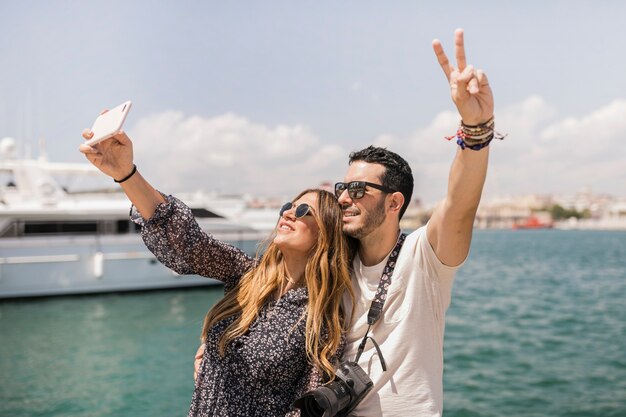 Pareja de turistas felices tomando selfie en celular contra el mar