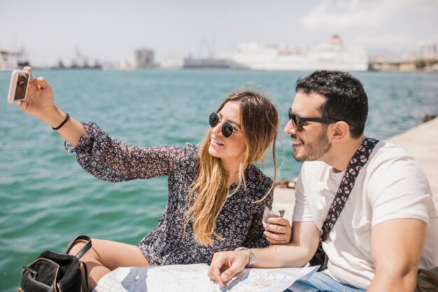 Pareja de turistas sentados con el mapa en el embarcadero tomando selfie en el teléfono celular