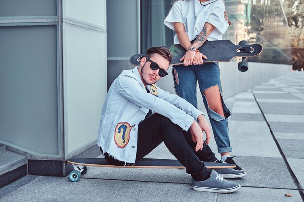 Pareja vestida de moda - joven hipster sentado en longboard y su novia de pie cerca del exterior.