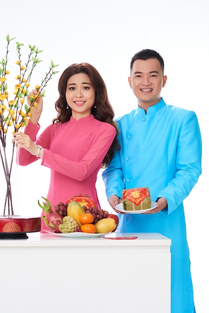 Pareja vietnamita en ropas tradicionales brillantes posando en estudio con frutas y flores.