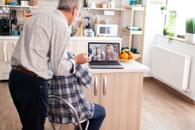 Las parejas ancianas durante la videoconferencia con la hija en la cocina usando la computadora portátil. Abuelos entusiastas hablando con la familia en línea usando la cámara web durante la discusión virtual, la comunicación moderna en línea