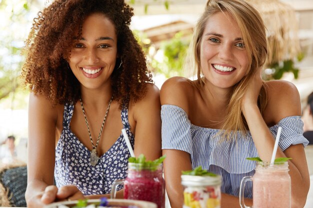 Las parejas de mujeres homosexuales tienen expresiones positivas, se sientan juntos en la cafetería, sonríen con alegría, disfrutan de deliciosos postres en la cafetería al aire libre. lesbianas multiétnicas hablan entre ellas. concepto de amor