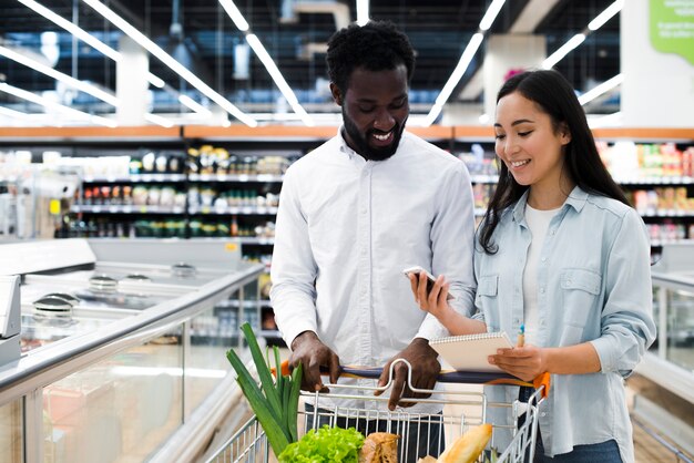 Pares alegres con el carro de la compra que comprueba en lista de compras móvil en el supermercado