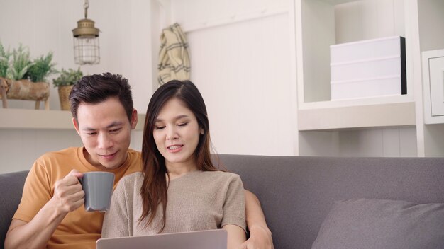 Los pares asiáticos que usan el ordenador portátil y beben la taza de café caliente en la sala de estar en casa, el par dulce goza