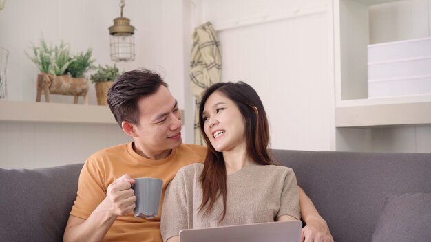 Pares asiáticos que usan el ordenador portátil y bebiendo la taza de café caliente en sala de estar en casa