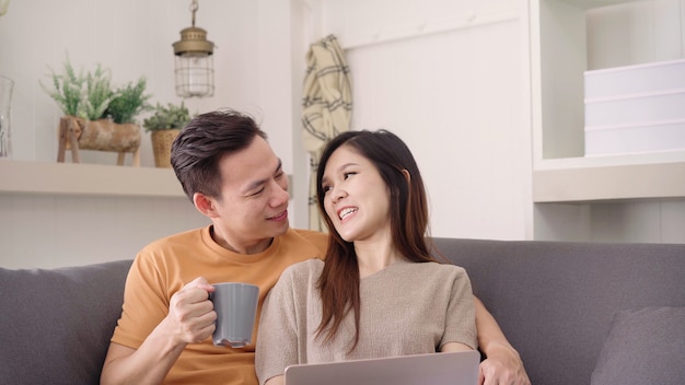 Pares asiáticos que usan el ordenador portátil y bebiendo la taza de café caliente en sala de estar en casa