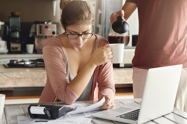 Pares caucásicos jovenes que tienen problemas financieros. Mujer estresada en vasos tomando café mientras administra el presupuesto familiar, sentada en la mesa de la cocina con documentos, computadora portátil y calculadora