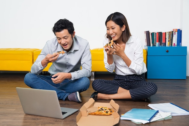 Pares felices asiáticos comiendo pizza mirando portátil