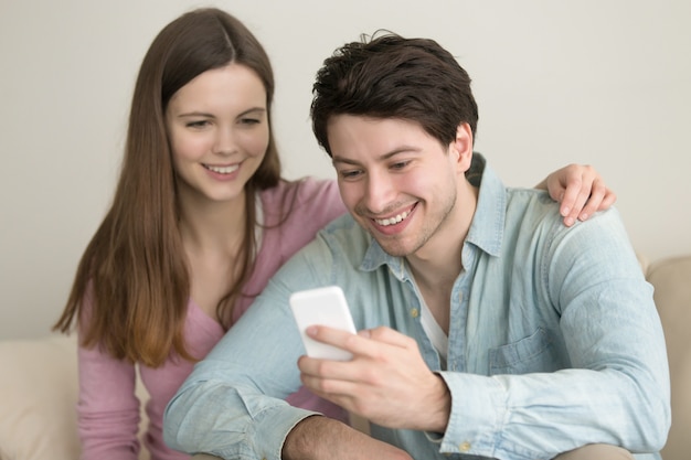Pares felices jovenes que se sientan en el sofá en casa usando smartphone
