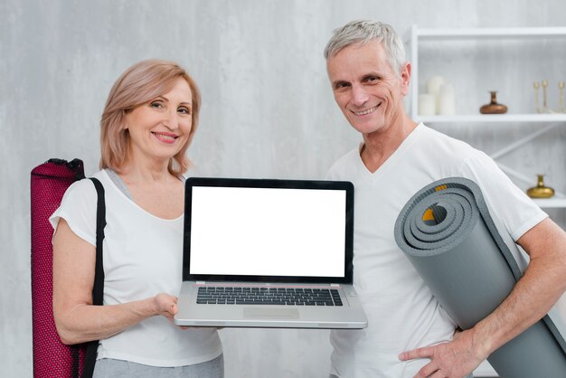 Pares felices que sostienen la estera y el ordenador portátil de la yoga con la pantalla blanca en casa