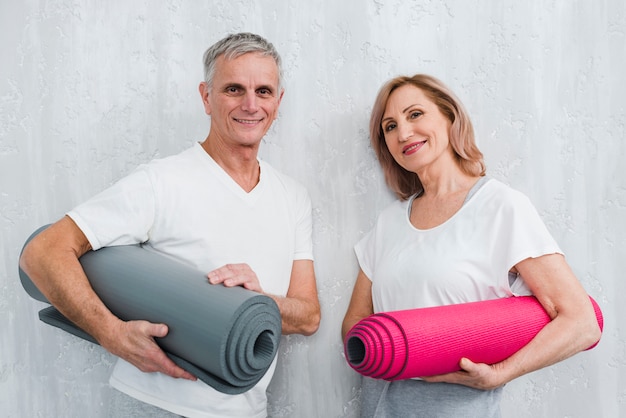 Pares felices que sostienen la pared blanca que se inclina que sostiene la estera rodada de la yoga