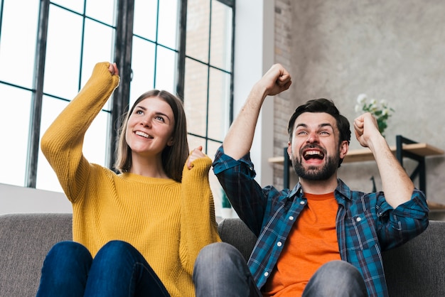 Foto gratuita pares jovenes emocionados que se sientan en el sofá que anima con alegría en casa