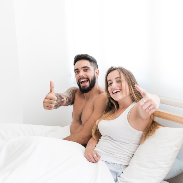 Foto gratuita pares jovenes que se sientan en la cama que gesticula los pulgares para arriba