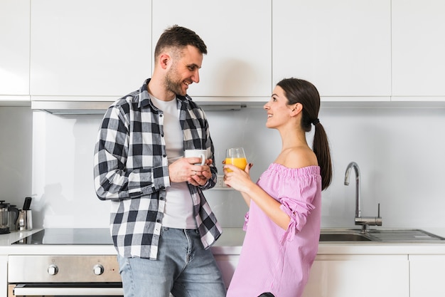 Pares jovenes sonrientes que sostienen la taza de café y el vidrio del jugo que se colocan en la cocina que mira la cámara