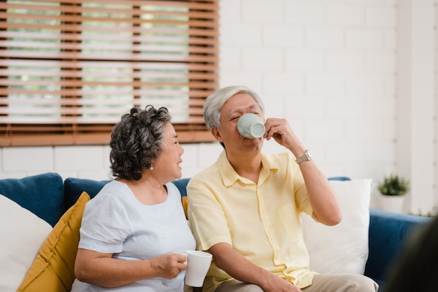 Los pares mayores asiáticos que beben el café caliente y que hablan juntos en sala de estar en casa, los pares disfrutan del momento del amor mientras que mienten en el sofá cuando está relajado en casa.