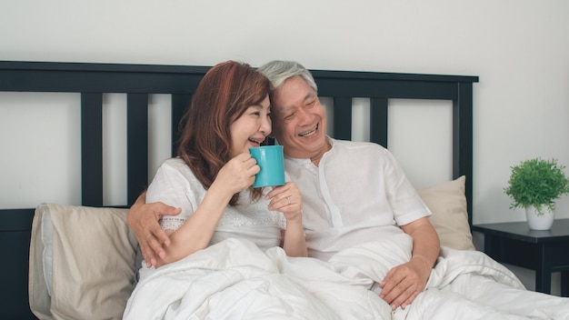 Pares mayores asiáticos que hablan en cama en casa. Los abuelos, el marido y la esposa chinos mayores asiáticos felices beben el café después de despertarse mientras que miente en cama en dormitorio en casa en el concepto de la mañana.