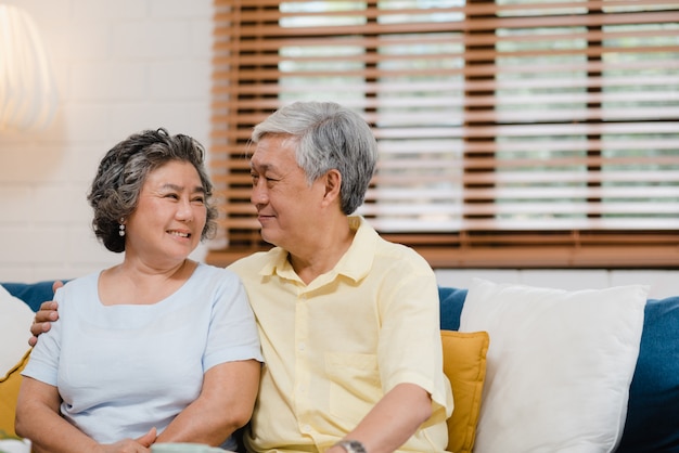 Los pares mayores asiáticos que llevan a cabo sus manos mientras que toman juntos en sala de estar, los pares que sienten parte feliz y se apoyan que mienten en el sofá en casa.