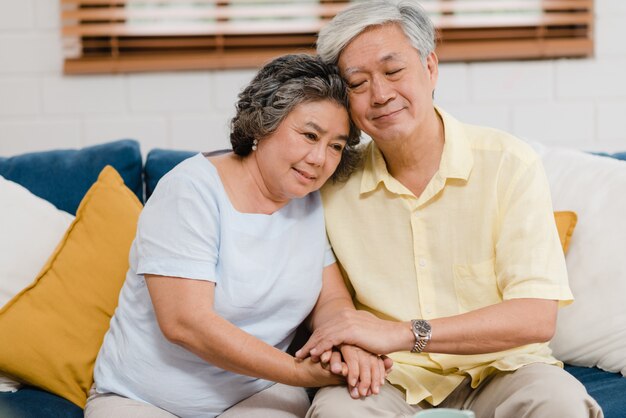 Los pares mayores asiáticos que llevan a cabo sus manos mientras que toman juntos en sala de estar, los pares que sienten parte feliz y se apoyan que mienten en el sofá en casa.