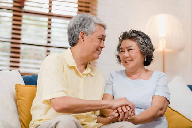 Los pares mayores asiáticos que llevan a cabo sus manos mientras que toman juntos en sala de estar, los pares que sienten parte feliz y se apoyan que mienten en el sofá en casa.
