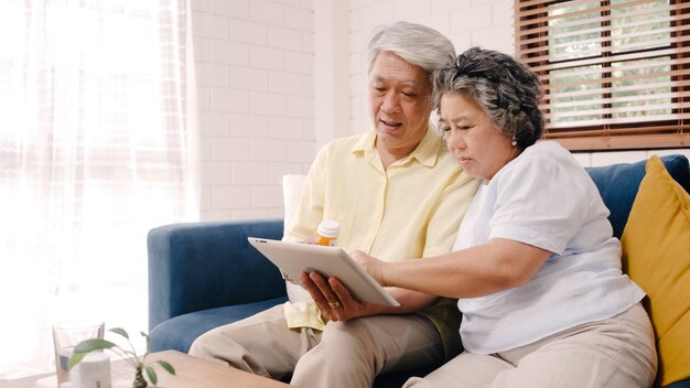 Pares mayores asiáticos que usan la información de la medicina de la búsqueda de la tableta en la sala de estar, pares que usan el tiempo junto mientras que miente en el sofá cuando está relajado en casa.