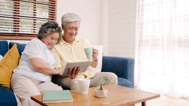 Los pares mayores asiáticos que usan la tableta y bebiendo café en la sala de estar en casa, los pares disfrutan del momento del amor mientras que mienten en el sofá cuando está relajado en casa.