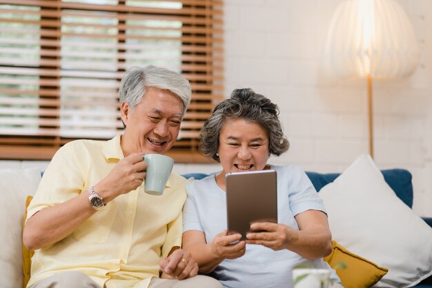 Los pares mayores asiáticos que usan la tableta y bebiendo café en la sala de estar en casa, los pares disfrutan del momento del amor mientras que mienten en el sofá cuando está relajado en casa.