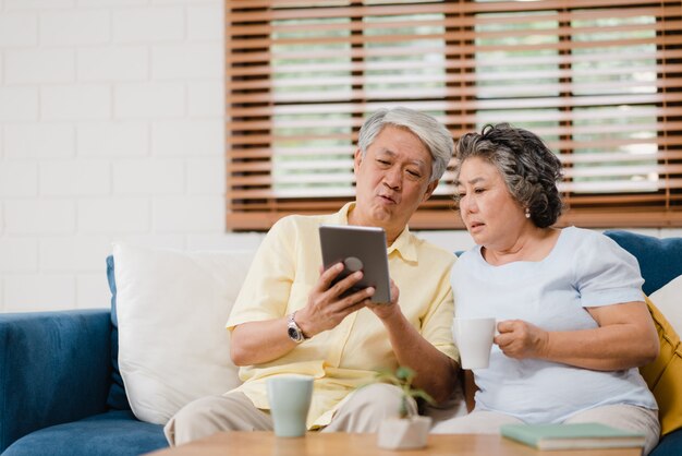 Los pares mayores asiáticos que usan la tableta y bebiendo café en la sala de estar en casa, los pares disfrutan del momento del amor mientras que mienten en el sofá cuando está relajado en casa.