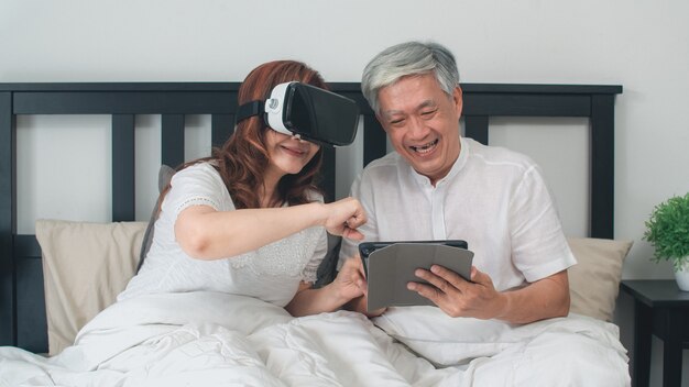 Pares mayores asiáticos que usan la tableta en casa. Abuelos chinos mayores asiáticos, sintiéndose diversión feliz y VR jugando juegos juntos mientras está acostado en la cama en el dormitorio en casa en el concepto de la mañana.