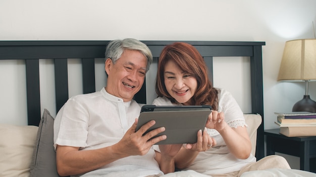 Pares mayores asiáticos que usan la tableta en casa. Los abuelos, marido y esposa chinos mayores asiáticos felices después de despertarse, viendo la película acostada en la cama en el dormitorio en casa en el concepto de mañana.