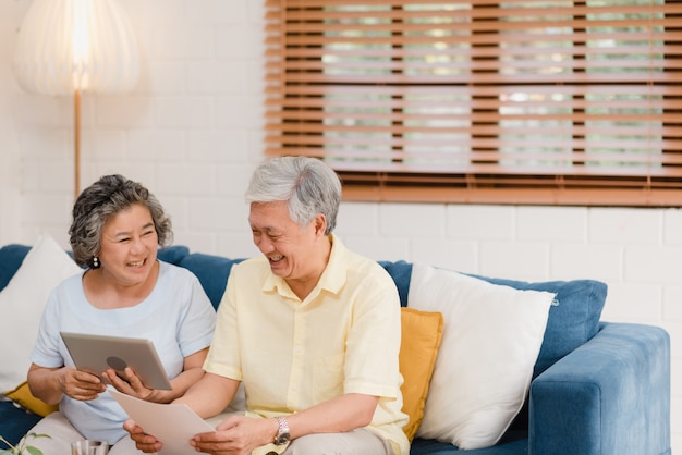 Los pares mayores asiáticos que usan la tableta que ve TV en sala de estar en casa, los pares disfrutan del momento del amor mientras que mienten en el sofá cuando está relajado en casa.