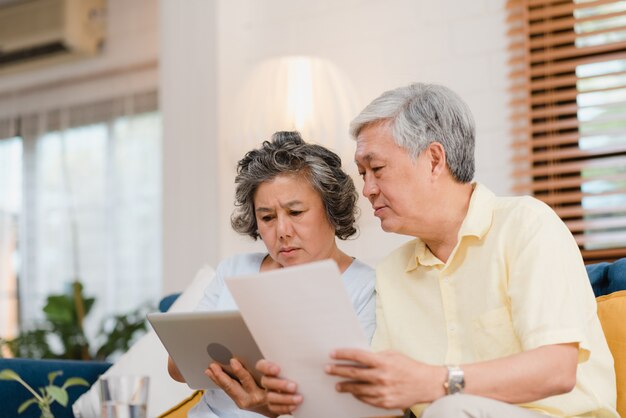 Los pares mayores asiáticos que usan la tableta que ve TV en sala de estar en casa, los pares disfrutan del momento del amor mientras que mienten en el sofá cuando está relajado en casa.