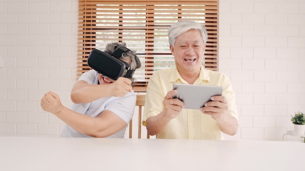 Pares mayores asiáticos que usan la tableta y el simulador de la realidad virtual que juegan a los juegos en la sala de estar, pares que sienten felices usando el tiempo junto que miente en la tabla en casa.