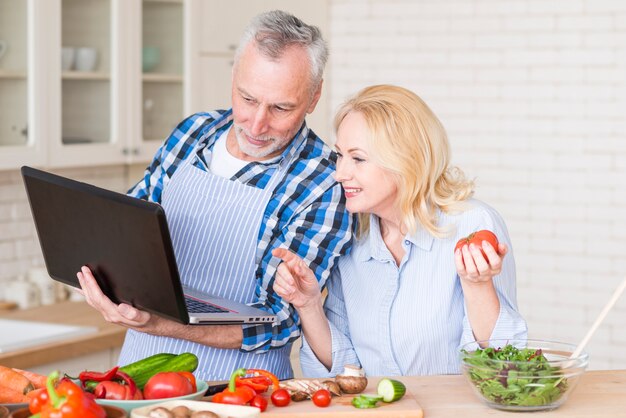 Pares mayores que miran el ordenador portátil mientras que prepara la ensalada vegetal en la tabla de madera