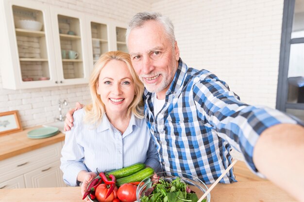 Pares mayores sonrientes que toman el autorretrato con verduras y el cuenco de ensalada en la cocina