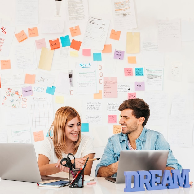 Foto gratuita pares sonrientes que se sientan en el escritorio con las computadoras portátiles contra la pared con las notas