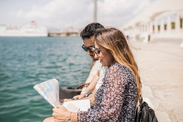 Pares turísticos sonrientes que se sientan en el embarcadero que mira el mapa