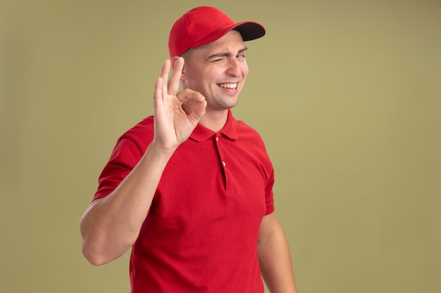 Parpadeó sonriente joven repartidor vestido con uniforme y gorra mostrando gesto bien aislado en la pared verde oliva con espacio de copia