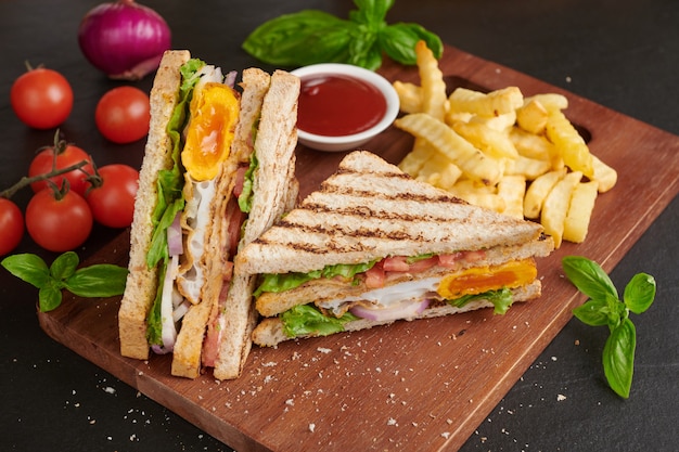 Foto gratuita a la parrilla y sándwich con tocino, huevo frito, tomate y lechuga servido en tabla de cortar de madera