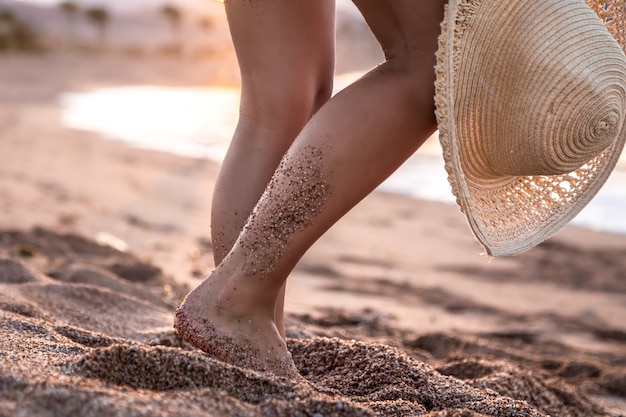 Parte del cuerpo. pies de una mujer de pie en la playa al atardecer con un sombrero.