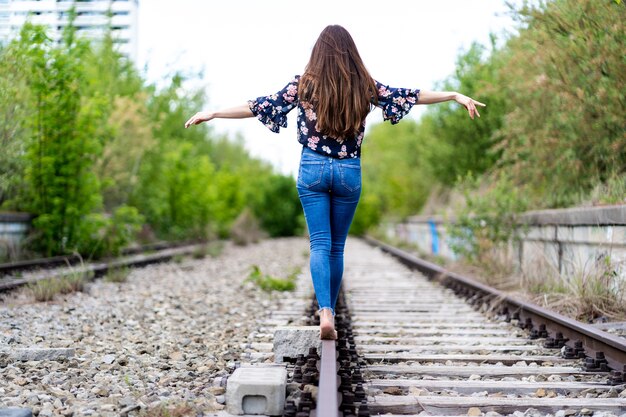 Parte posterior de una mujer joven caminando por los rieles del tren descalzo y tratando de mantener el equilibrio