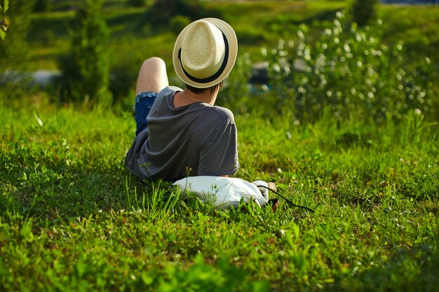 La parte trasera del joven y atractivo hombre con estilo moderno en ropa casual con sombrero en gafas sentado en el parque en la hierba verde