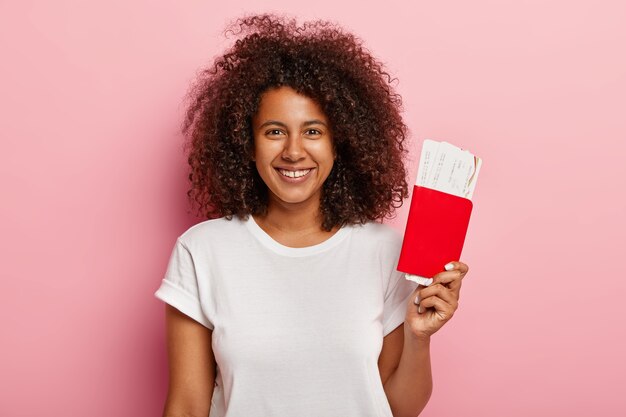 La pasajera positiva tiene boletos de embarque y pasaporte, se prepara para un vuelo, disfruta viajar, tiene un peinado rizado, usa una camiseta blanca, sonríe ampliamente con dientes perfectos