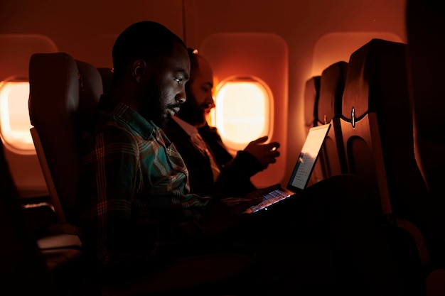Pasajero independiente trabajando en una laptop durante la puesta de sol, volando en avión en un vuelo comercial con servicio de aerolínea internacional. Turista masculino que viaja por trabajo o vacaciones.