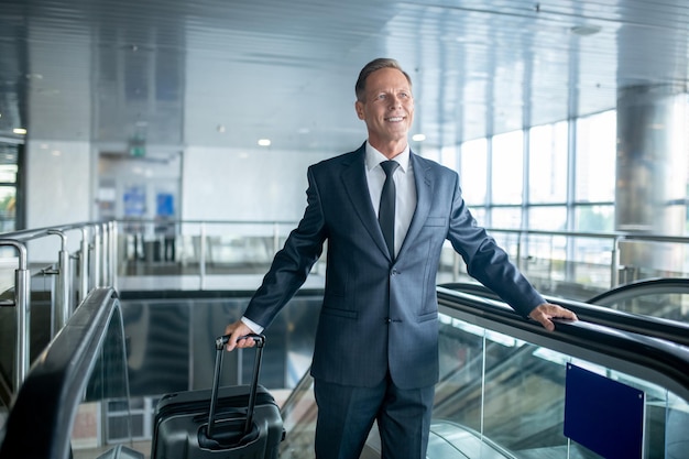 Pasajero masculino de gran espíritu con la maleta con ruedas bajando de la escalera móvil en el aeropuerto