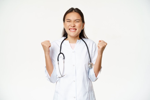 Pasante asiático feliz celebrando. Médico médico triunfando y regocijándose, di que sí con la bomba de puños cerrados, de pie extático sobre fondo blanco.