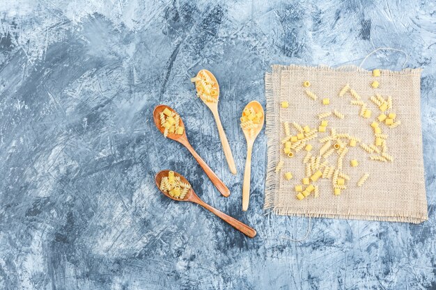 Pasta cruda en cucharas de madera sobre un yeso y un trozo de fondo de saco. vista superior.