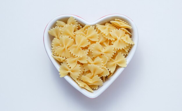 Pasta cruda farfalle en un recipiente en forma de corazón en la pared blanca, vista superior.